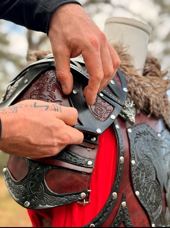 Hands fastening the buckled shoulder strap of an oxblood and black leather pauldron with carved knotwork, brass rivets, and a dragon concho