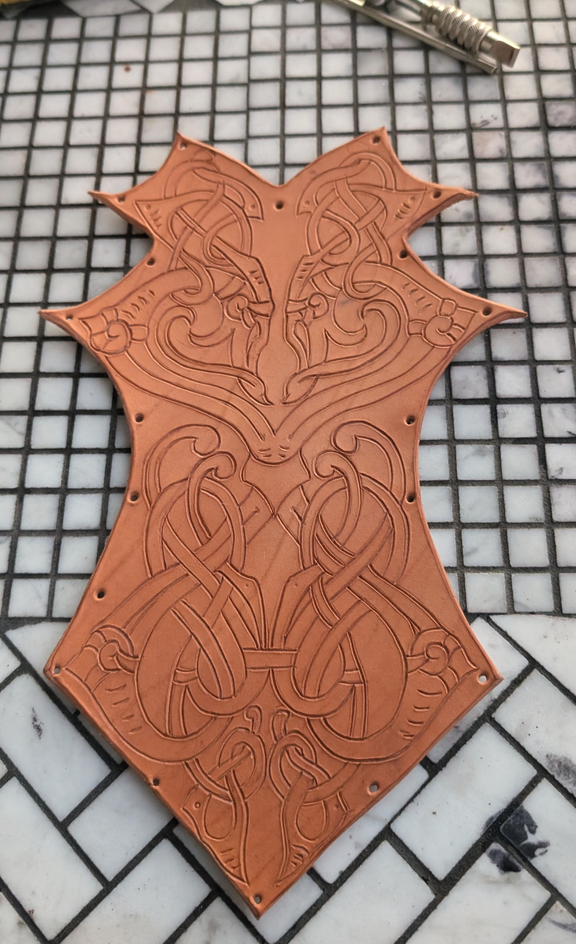 A freshly tooled leather chest panel with knotwork carved into undyed leather on a tiled work surface