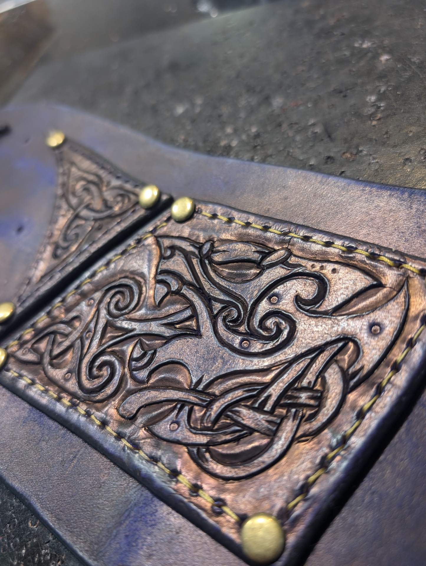Macro close-up of carved and burnished Celtic knotwork on a leather panel with brass rivets and yellow stitching