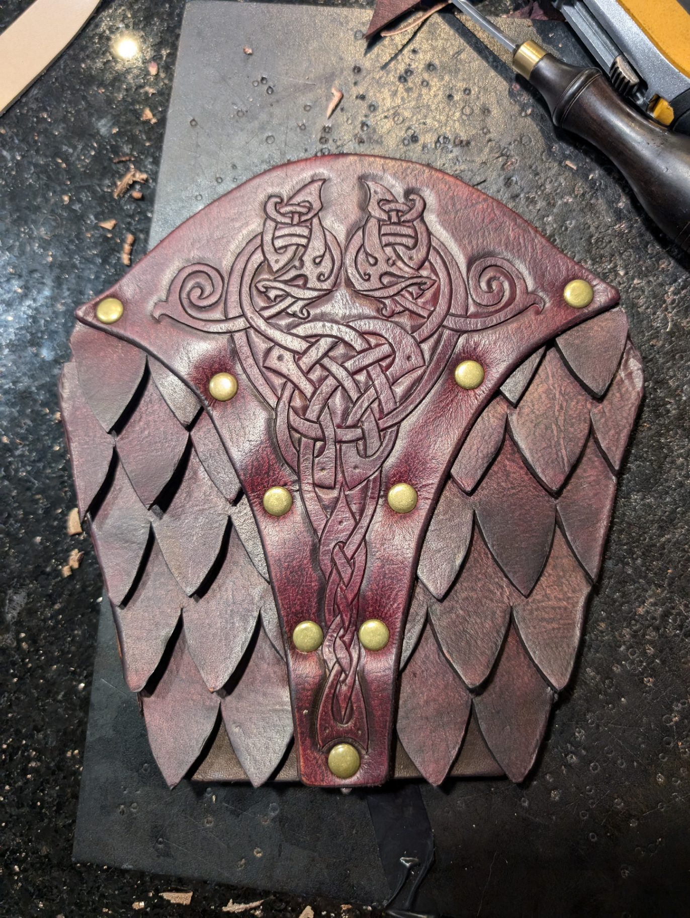 Oxblood leather scale pauldron with carved beast knotwork and overlapping leather scales secured by brass rivets
